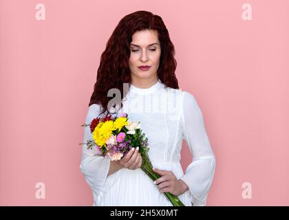 Femme regardant vers le bas tenant bouquet de fleurs avec fond rose.Belle femme pensive en robe blanche romantique tenant un bouquet de fleurs. Banque D'Images