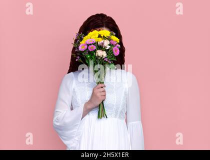 Femme avec bouquet de fleurs devant le visage sur fond rose.Portrait de studio de femme joueur tenant un bouquet de fleurs devant son visage. Banque D'Images