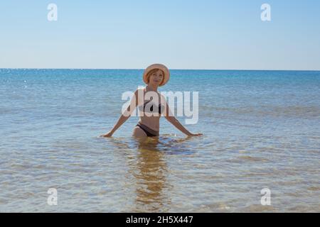 Une femme européenne en maillot de bain et en chapeau de taille dans la mer sourit et regarde dans la caméra.Vacances en bord de mer. Banque D'Images