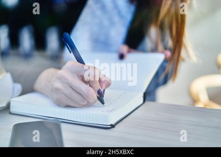 Par le verre anonyme femme entrepreneur assis à la table avec un smartphone et écrire des notes de travail dans le bloc-notes Banque D'Images