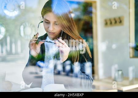 À travers le verre de la travailleuse concentrée prenant des lunettes tout en lisant des notes dans le bloc-notes à la table avec café et croissant dans le café Banque D'Images