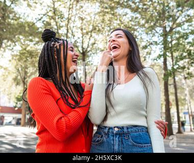 Joyeuses amies multiraciales dans des vêtements élégants debout et riant contre des arbres verts dans la rue à la lumière du jour Banque D'Images