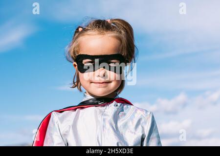 Mignon enfant portant le costume de super-héros mascarade et masque de l'œil debout contre le ciel bleu et regardant l'appareil photo Banque D'Images