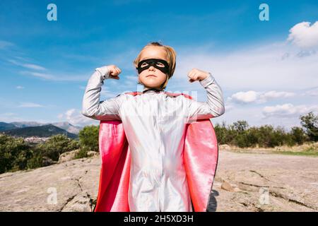 Une fille forte en cape super-héro qui a les mains pour montrer sa force pour protéger le monde tout en se tenant debout sur une colline et en regardant loin avec confiance Banque D'Images