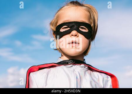 Mignon enfant portant le costume de super-héros mascarade et masque de l'œil debout contre le ciel bleu et regardant l'appareil photo Banque D'Images