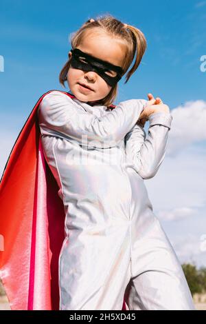 D'en-dessous, un enfant mignon portant un costume de super-héros mascarade et un masque pour les yeux debout contre le ciel bleu et regardant l'appareil photo Banque D'Images