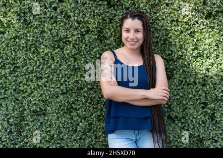 Positive hispanique femelle avec tatouage et long noir tressé cheveux souriant et regardant la caméra tout en se tenant près de plantes vertes Banque D'Images