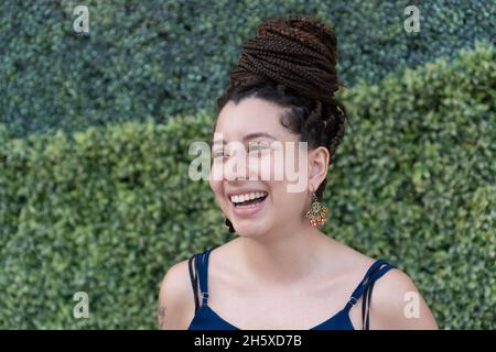 Positive hispanique femelle avec tatouage et long noir tressé cheveux souriant et regardant la caméra tout en se tenant près de plantes vertes Banque D'Images