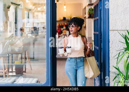 Jeune Afro-américaine positive avec des cheveux bouclés dans des vêtements décontractés tout en laissant un café moderne avec des tasses à café à emporter et des sacs en papier Banque D'Images