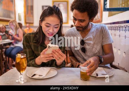 Une jeune femme asiatique montrant des photos sur un smartphone à un petit ami ethnique assis à table au restaurant Banque D'Images
