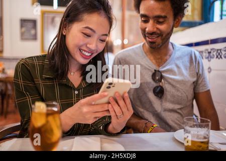 Une jeune femme asiatique montrant des photos sur un smartphone à un petit ami ethnique assis à table au restaurant Banque D'Images