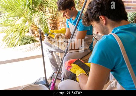 Corps complet de jeunes hommes professionnels de nettoyage en uniforme et gants utilisant des téléphones mobiles tout en se reposant sur un canapé confortable sur la terrasse de la cotta moderne Banque D'Images