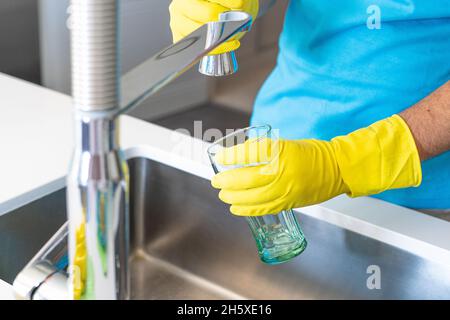 Récolte anonyme mâle dans des gants en caoutchouc jaune remplissant le verre avec de l'eau du robinet pendant la routine de nettoyage à la maison Banque D'Images