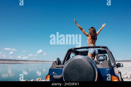 Vue arrière d'une jeune femme en haut et d'un short en denim, les bras relevés dans une voiture de sport décapotable garée sur un sable fin Banque D'Images