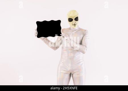 Personne vêtue d'un costume argenté et d'un masque vert étranger, tenant un tableau noir, sur fond blanc Banque D'Images