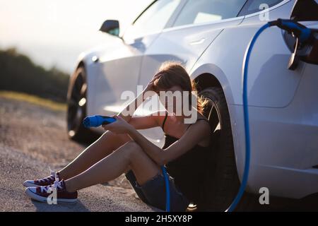 Belle jeune fille triste à côté d'une voiture électrique. Tenant un câble de charge. Rétroéclairage du coucher du soleil. Banque D'Images