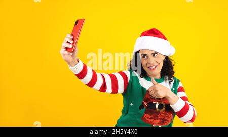 Portrait d'une femme latine senior tenant un téléphone mobile prenant un selfie photo dans un concept de noël sur fond jaune Banque D'Images