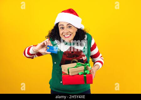 Portrait d'une femme adulte latine tenant une carte de crédit pour faire du shopping à Noël portant un chapeau de père noël sur fond jaune au Mexique en Amérique latine Banque D'Images