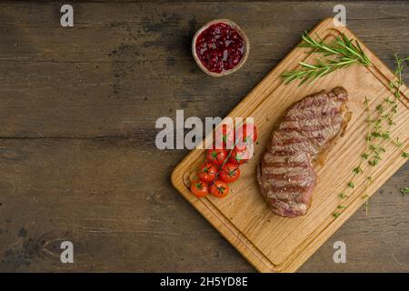 Steak de stripine grillé avec tomates cerises, thym, romarin et sauce aux baies de lingonis sur planche à découper en bois.Délicieuse viande juteuse et tendre.Horizont Banque D'Images