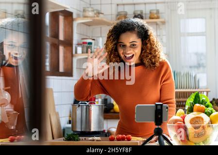 Jeune Afro-américaine regardant le téléphone mobile et de dire salut sur les médias sociaux dans la cuisine à la maison, concept de cours de cuisine virtuelle Banque D'Images