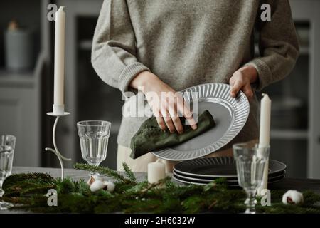 Gros plan de la jeune femme qui a mis des assiettes sur la table à manger décorée pour Noël avec des branches de sapin et des bougies dans des tons gris Banque D'Images