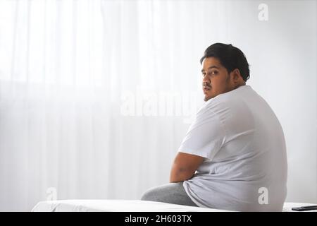 Un gros homme assis avec une expression sérieuse sur le visage dans sa chambre. Banque D'Images
