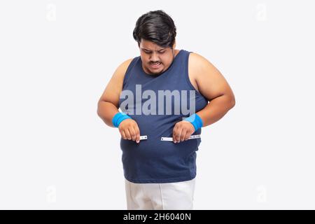 Portrait d'un gros homme essayant d'envelopper le ruban de mesure autour de sa taille sur fond simple. Banque D'Images