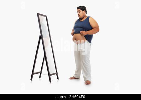 Portrait d'un gros homme regardant le bellyfat dans le miroir avec surprise sur fond simple. Banque D'Images