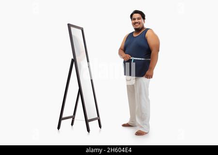 Portrait d'un gros homme souriant avec du ruban de mesure autour de sa taille et un miroir sur fond Uni. Banque D'Images