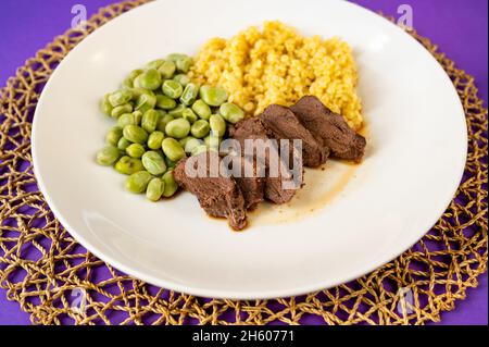 Viande de cerf maigre en tranches, pâtes cuites aux haricots et semoule « tarhona » sur l'assiette sur la plaque de bambou circulaire sur la table, en gros plan. Banque D'Images