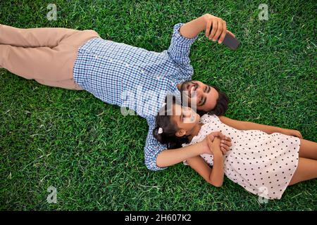 UN PÈRE ET UNE FILLE QUI CLIQUENT SUR UN SELFIE MOBILE TOUT EN SE RELAXANT SUR L'HERBE Banque D'Images