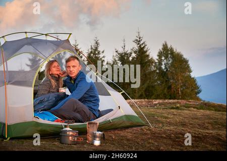 Femme randonneur tenant une tasse de café, assise à côté de son mari dans une tente touristique.Couple heureux voyageurs se reposant dans une tente de camp avec beau ciel sur fond tout en randonnée ensemble dans les montagnes. Banque D'Images