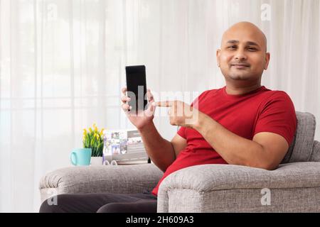 Un homme chauve pointant vers son téléphone mobile assis sur un canapé dans sa chambre. Banque D'Images