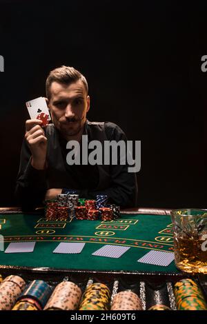 Beau joueur de poker avec deux as dans ses mains et des jetons assis à la table de poker sur fond noir Banque D'Images