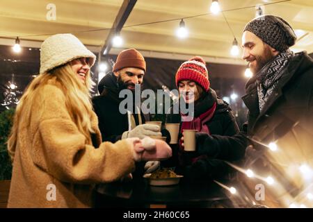 Des amis souriants avec des tasses de vin chaud s'amusent, passant du temps ensemble à la foire d'hiver le soir.Vacances, concept de Noël Banque D'Images