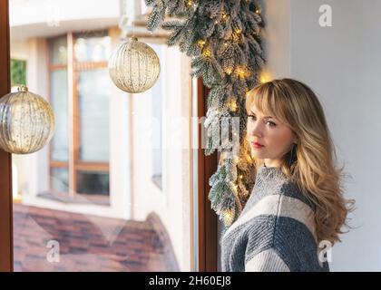 Portrait d'une belle femme blonde portant un pull chaud.Femme attirante adulte à la maison pose par la fenêtre décorée avec des ornements de Noël.FEMA Banque D'Images