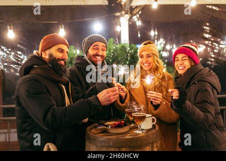 Des amis souriants avec des tasses de vin chaud s'amusent, passant du temps ensemble à la foire d'hiver le soir.Vacances, concept de Noël Banque D'Images