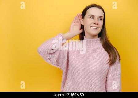 Joyeux curieux nouveau nez souriant jeune femme caucasienne 20s vêtu d'un chandail tricoté rose, essayez de vous entendre écouter attentivement Banque D'Images