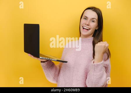 Souriant rédacteur pigiste positive jeune femme tenir ordinateur portable pc chat ne gagnant de la gestuelle de serrage, habillé en tricot rose chandail Banque D'Images
