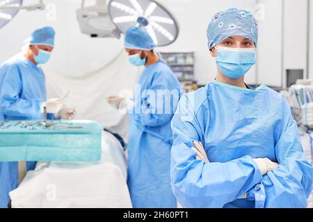 Jeune médecin ou infirmière en salle d'opération avec bras croisés dans la salle d'opération avec l'équipe de chirurgie et le patient Banque D'Images