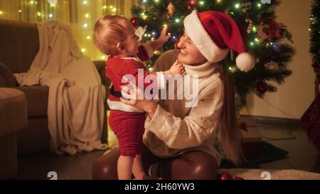 Mignon petit garçon portant le costume du Père Noël assis avec la mère à côté de l'arbre de Noël.Familles et enfants célébrant les vacances d'hiver. Banque D'Images