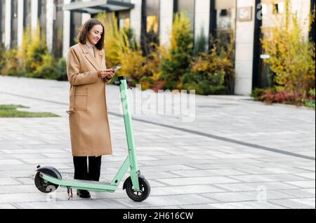 Femme heureuse en manteau d'automne utilisant un smartphone pour louer un scooter électrique moderne avec une application, lit le code qr avec une caméra de téléphone mobile.Transport urbain alternatif Banque D'Images