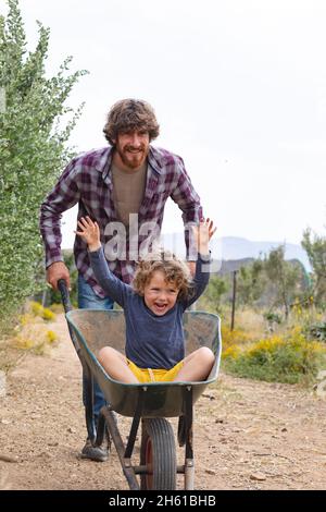Jeune homme poussant un fils joyeux et excité assis avec les mains soulevées dans la brouette sur la passerelle Banque D'Images