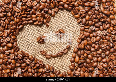 Vue d'un cadre incliné en forme de cœur fait de grains de café torréfiés avec un smiley sur une toile de sackCloth. Banque D'Images