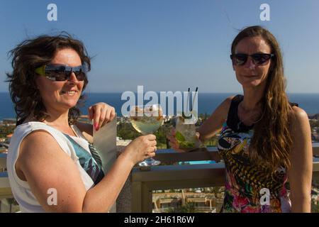 Des femmes ravie avec des cocktails sur la terrasse sur le toit.Des amis souriants avec des lunettes de soleil pour fêter la fête le jour ensoleillé.Les jeunes femmes s'amusent Banque D'Images