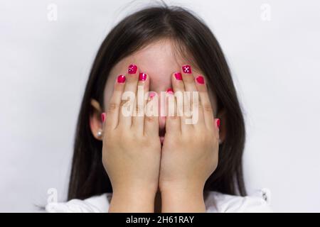 Timide petite fille couvre son visage avec les mains montrant les ongles vernis rose sur fond blanc Banque D'Images