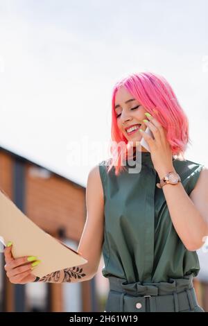 femme d'affaires ravie avec des dossiers de maintien de cheveux roses et de parler sur le smartphone à l'extérieur Banque D'Images