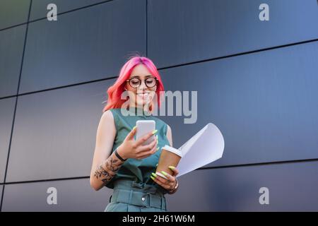 femme d'affaires heureuse et tatouée dans des écouteurs utilisant un smartphone tout en tenant une tasse en papier Banque D'Images