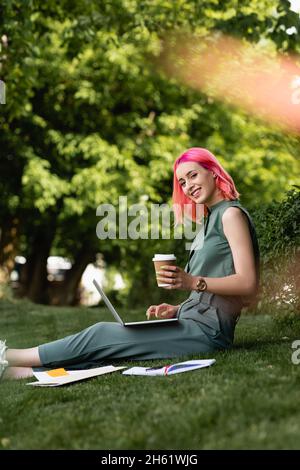 une femme heureuse avec des cheveux roses tenant une tasse en papier et utilisant un ordinateur portable sur l'herbe Banque D'Images