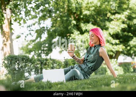 jeune femme gaie avec des cheveux roses tenant une tasse de papier près d'un ordinateur portable sur l'herbe Banque D'Images